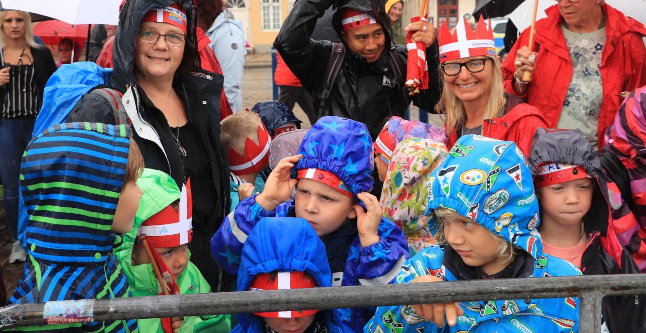 “Det er en kæmpe ære, at dronningen kommer og besøger os fra mindretallet”. Begejstringen hos Christina Strunz-Ewel, dagsinstitutionsleder i Lyksborg Børnehave, er ikke til at tage fejl af. På trods af at regnen vælter ned over Flensborg Havn, smiler hun stort. Rundt omkring hende står en gruppe børnehavebørn, der alle bærer dannebrogskroner med teksten “Velkommen til Sydslesvig” under regnjakkehætten. “Vi har de seneste uger undervist børnene i grænser, mindretal og historie forud for jubilæet for grænsedr