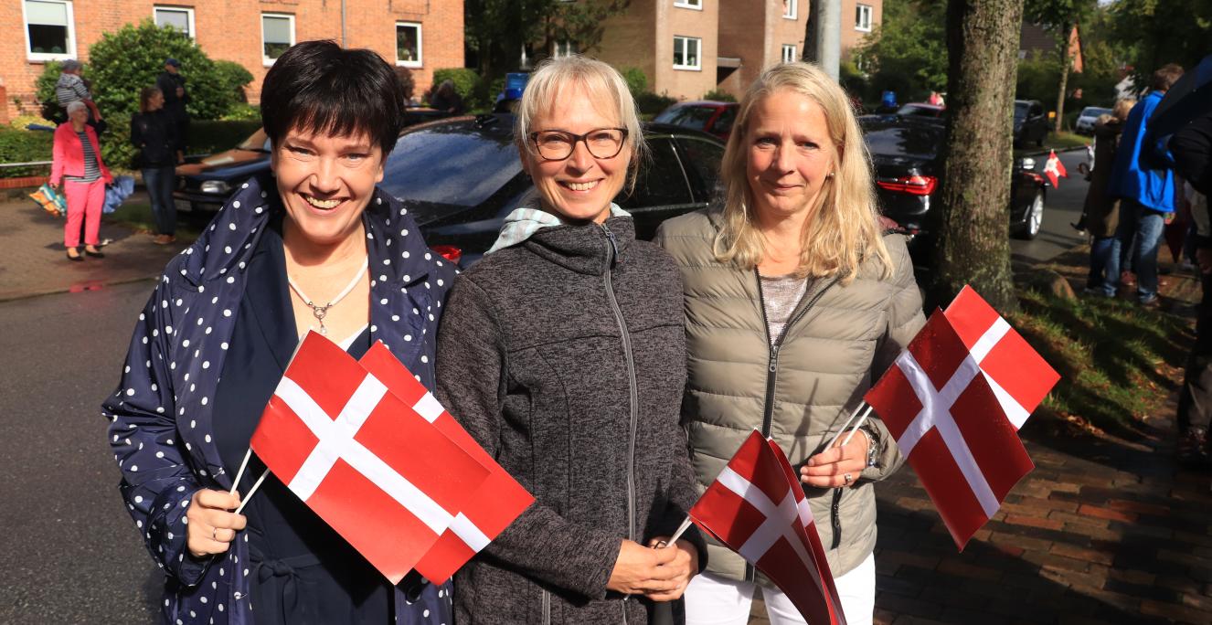 Nerongsallee, en ganske almindelig villavej i det vestlige Flensborg, er afspærret. Foran det danske alderdomshjem, der har ligget på adressen siden 1950, har en mindre menneskeskare samlet sig. En børnehave øver sig i at råbe “hurra” så højt, de kan. Men få minutter efter, da dronningen er ankommet, glemmer næsten halvdelen af børnene at råbe noget som helst. De er alt for betagede af dronningen og hendes følge. Foran alderdomshjemmet står også Gitte Krugmann (i midten) og Sonja Schilling-Lenz (th.), der e